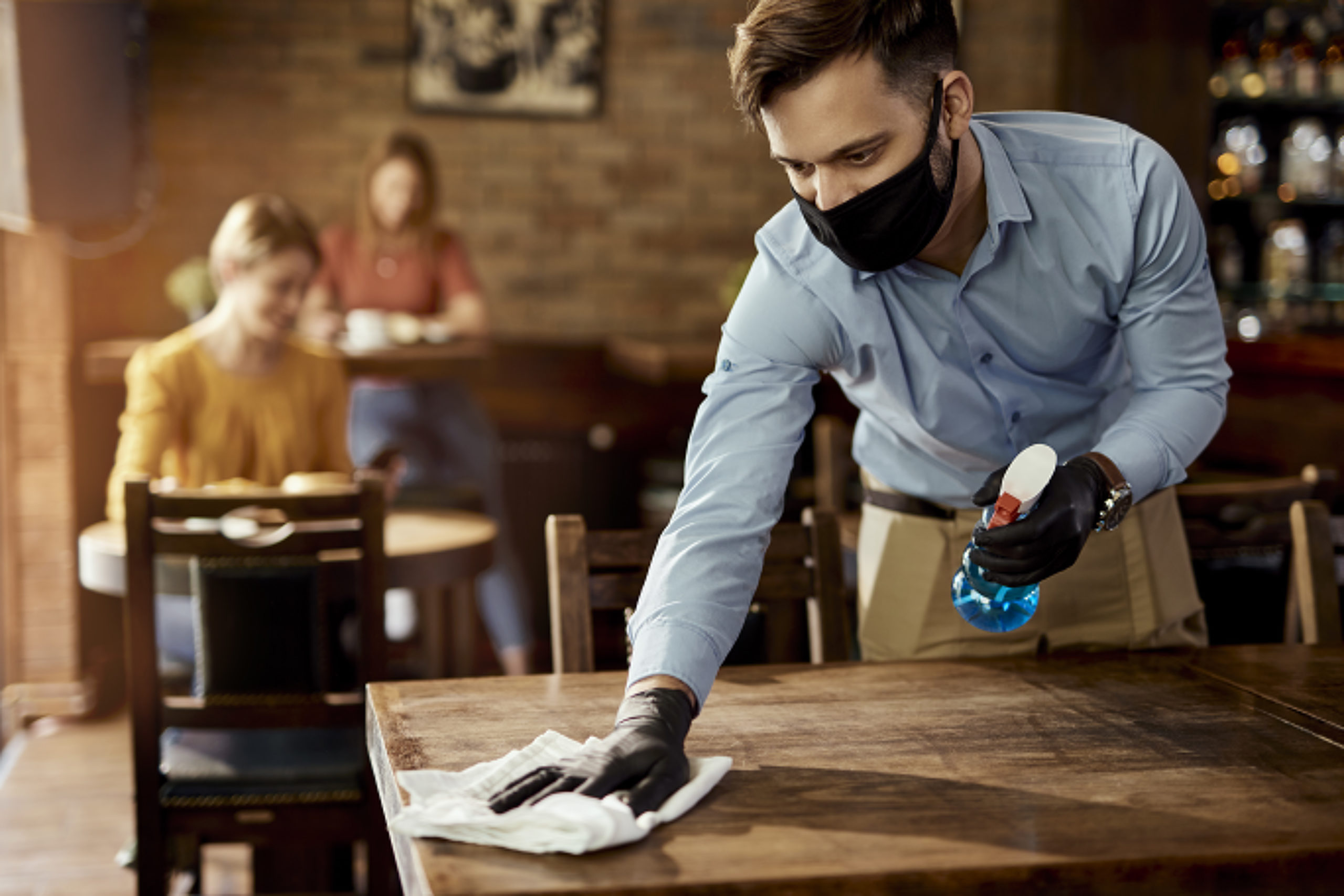 Waiter with protective face mask disinfecting tables in a pub.