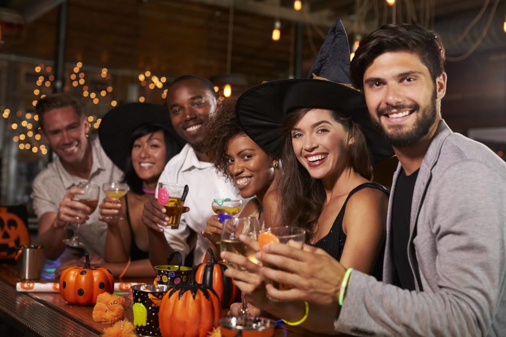 Mixed group of friends in Halloween costumes raising glasses at a bar and smiling.