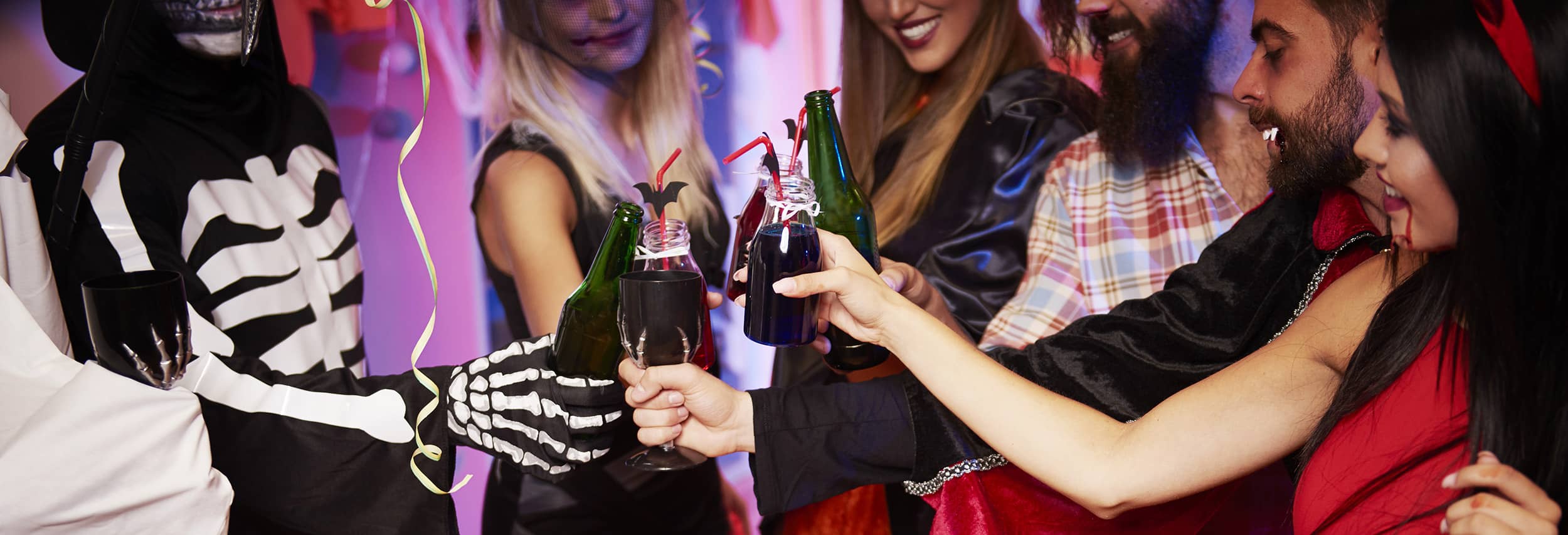 Closeup of several women dressed in Halloween costumes toasting mixed drinks and beers at a Halloween event.