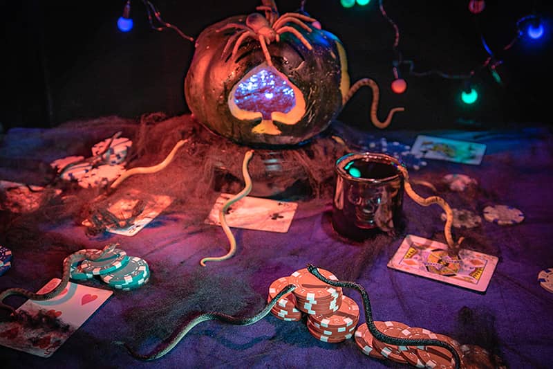 Picture of a jack-o-lantern on a table with snakes, bugs, cards, and poker chips giving a Halloween gaming feeling.