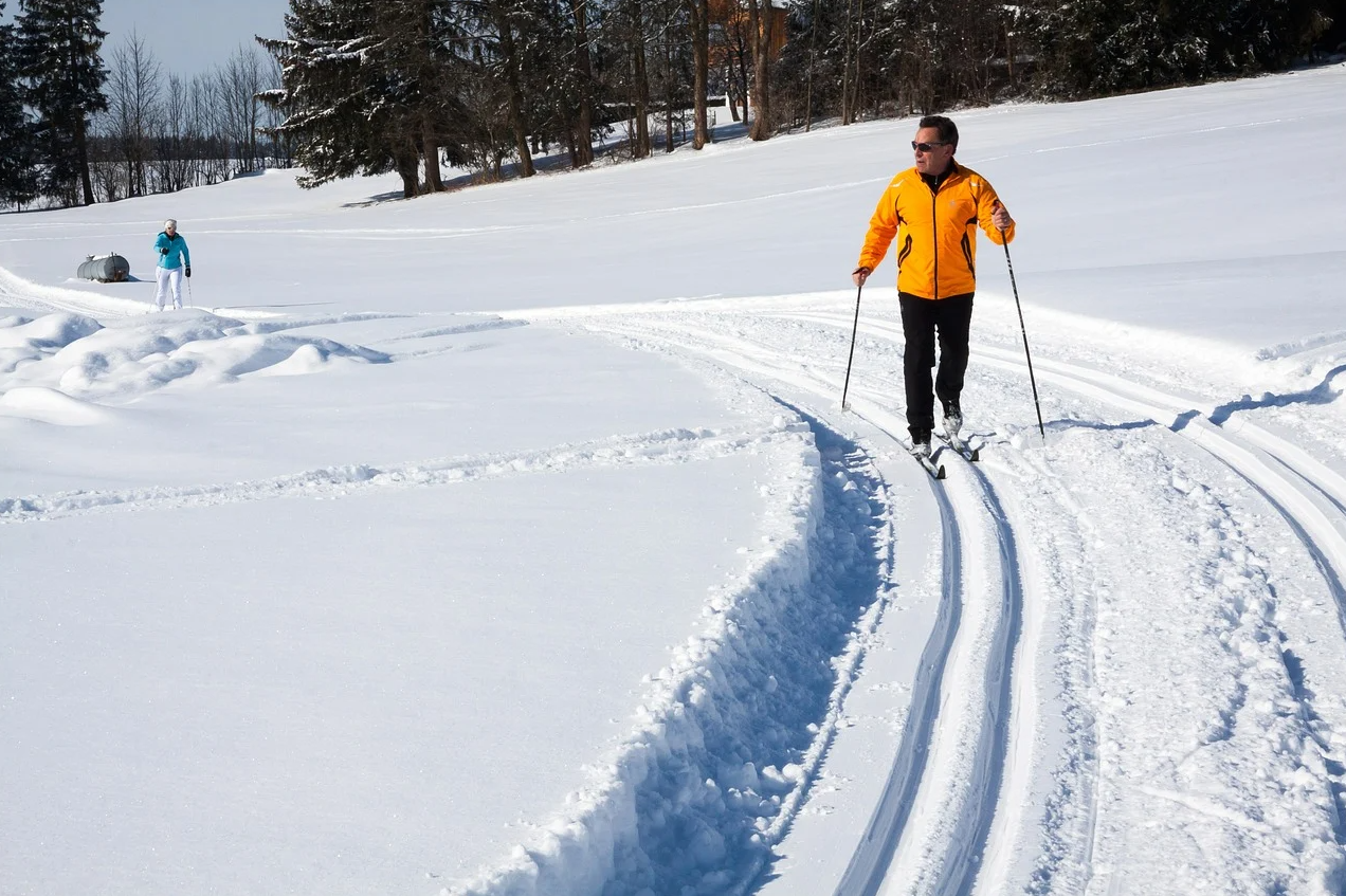 A man in a yellow jacket is leading on a curved cross-country track through the snow as in the distance, a woman in a teal jacket follows. 