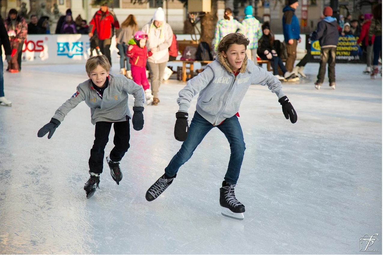 Two boys in jeans, gray sweatshirts and ice skates race across an outdoor rink on a sunny afternoon