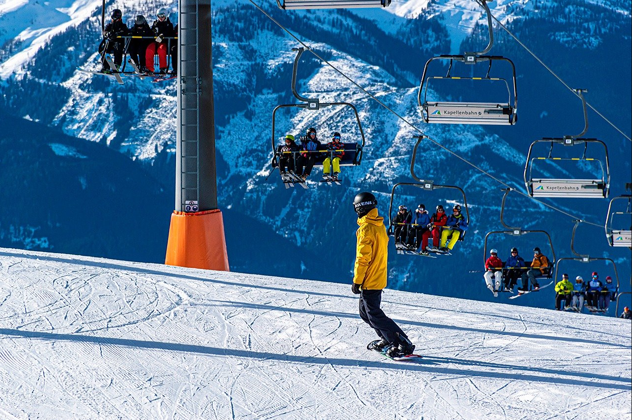 On a bright and sunny day, a ski lift is carrying colorfully clothed skiers to the top of a snow-covered mountain while a snowboarder with a helmet and yellow jacket skis down