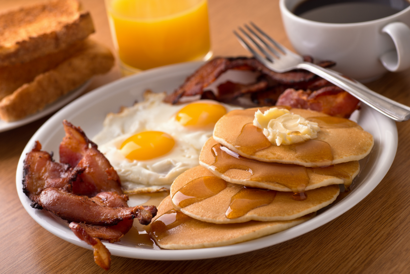 A breakfast plate loaded with pancakes, eggs over easy and bacon sits next to a glass of orange juice