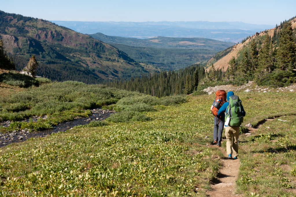 A couple with their backs facing the viewer hike down a trail with mountains in the distance planning a summer trip to cripple creek