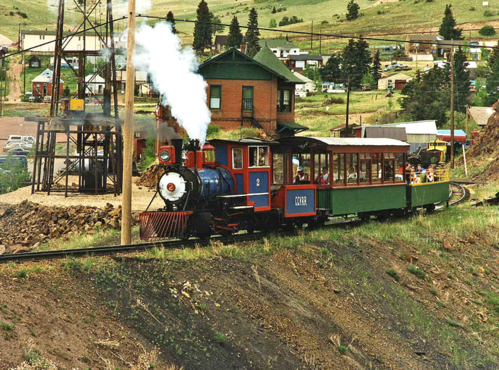 A steam powered engine pulls an open air green and red train on a track atop a steep hill with historic brick buildings in the background