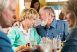A mature couple, the woman with blonde short hair and the man with white short hair and a beard, share an intimate moment smiling at a table filled with food and drinks