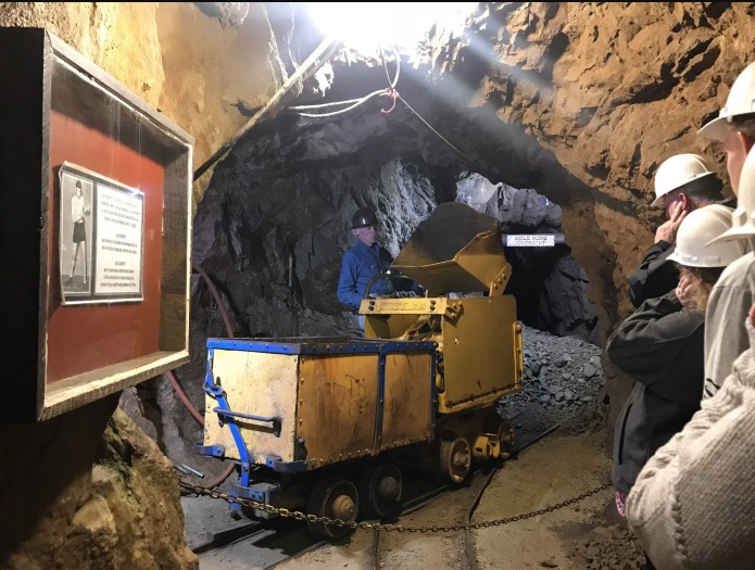 A picture of several people in a man-carved cavern in the earth viewing a yellow mini hauler of rocks in the Mollie Kathleen Mine