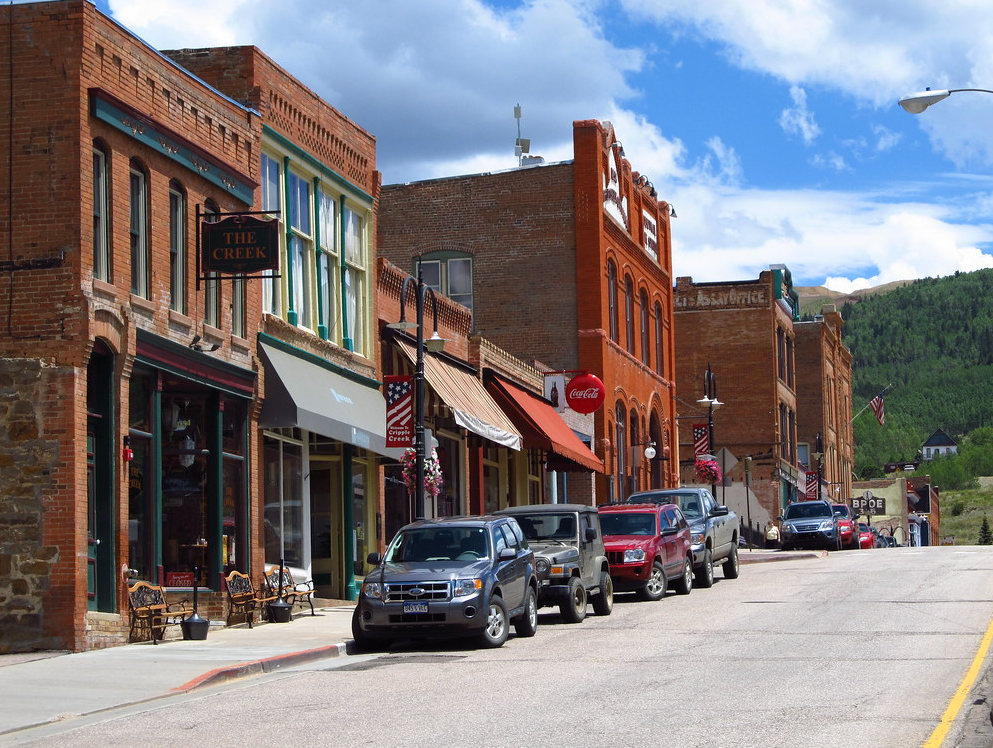 A view down the historic buildings lining main street in Cripple Creek Colorado with cars parked in front on a sunny day.