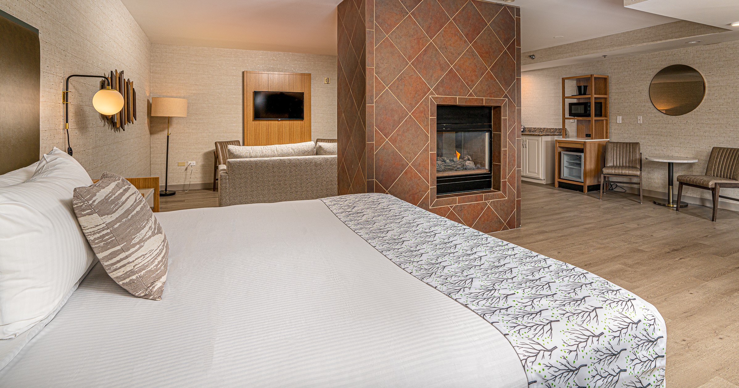 A king sized bed is neatly made in a beautifully decorated neutral hotel room with a brick fireplace.