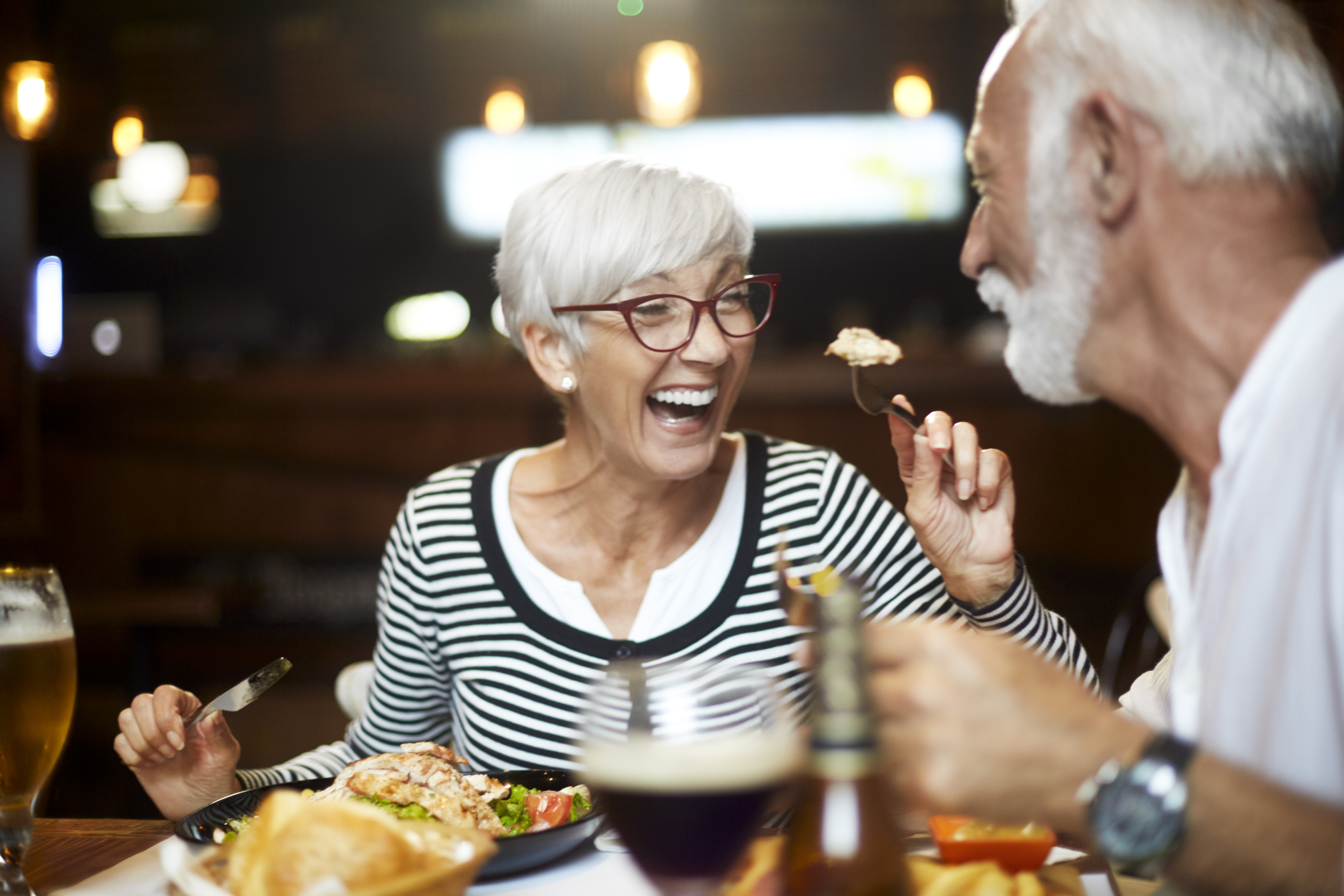 Elderly couple cheerfully feeding each other during a meal in a restaurant.