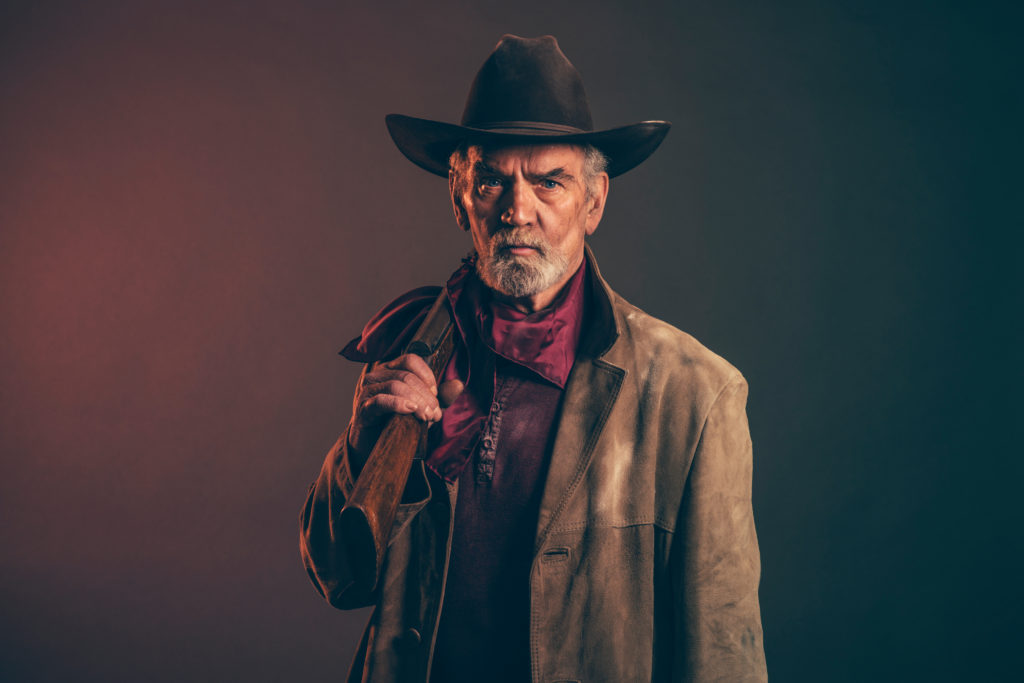 Old rough western cowboy with gray beard and brown hat holding rifle. Low key studio shot.